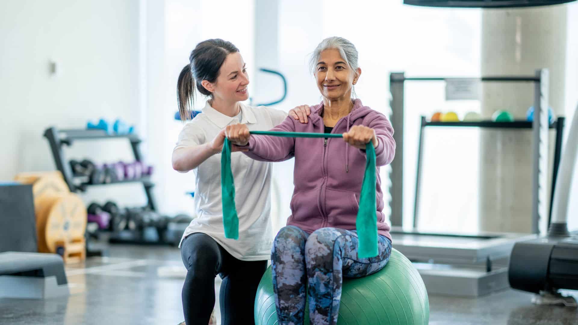 A woman assists an older woman as she balances on an exercise ball